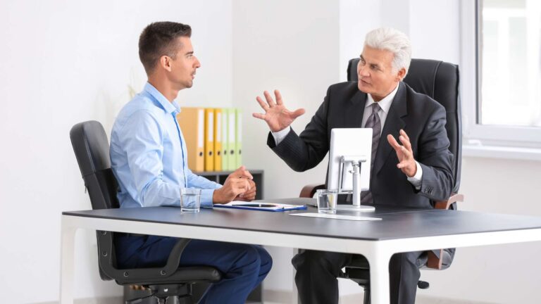 Two businessmen sit across from each other at a desk in a modern office, one speaking with animated hand gestures as the other listens attentively.
